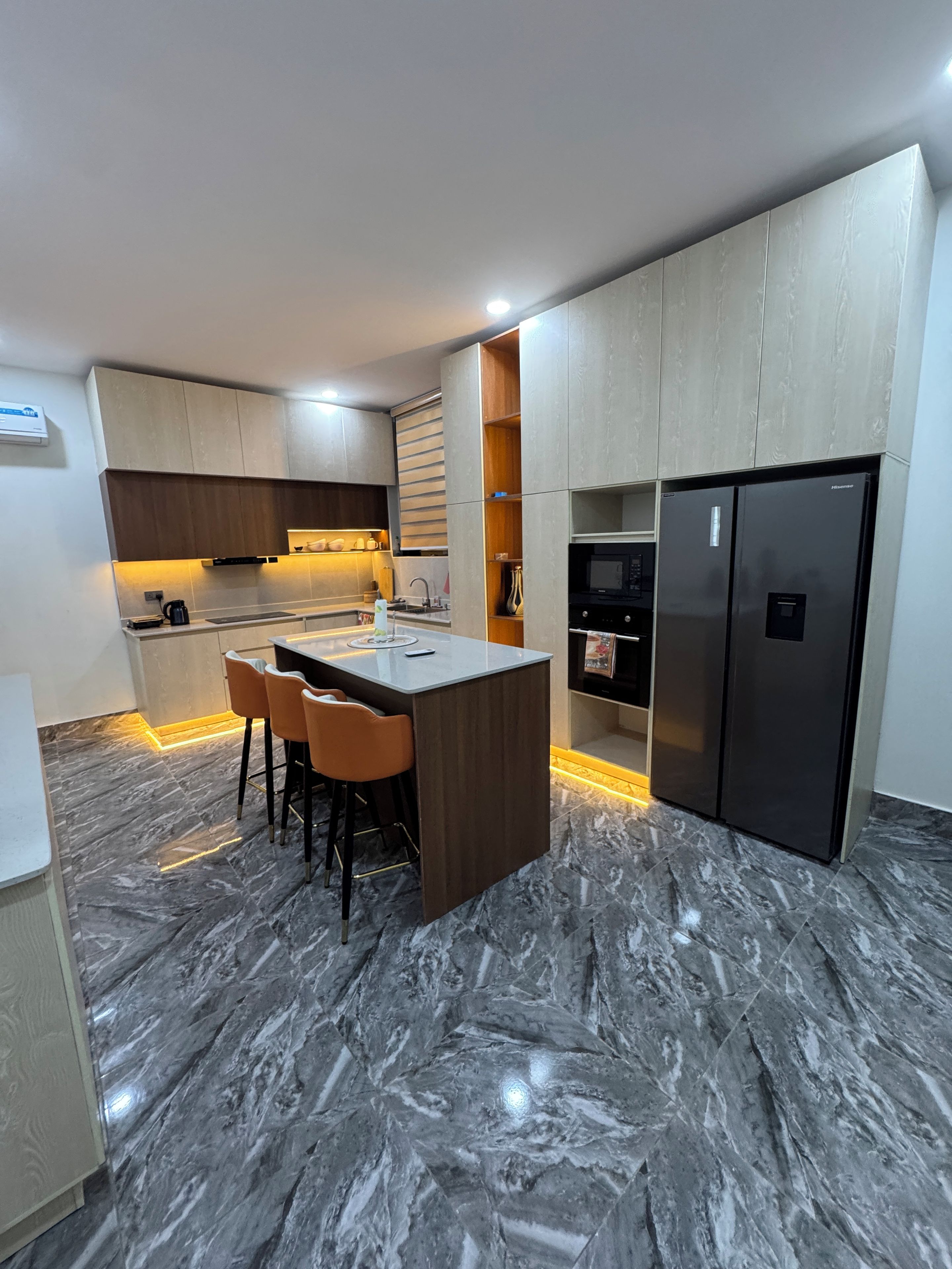A modern kitchen with a marble floor, a central island with bar stools, built-in appliances, and wooden cabinetry.
