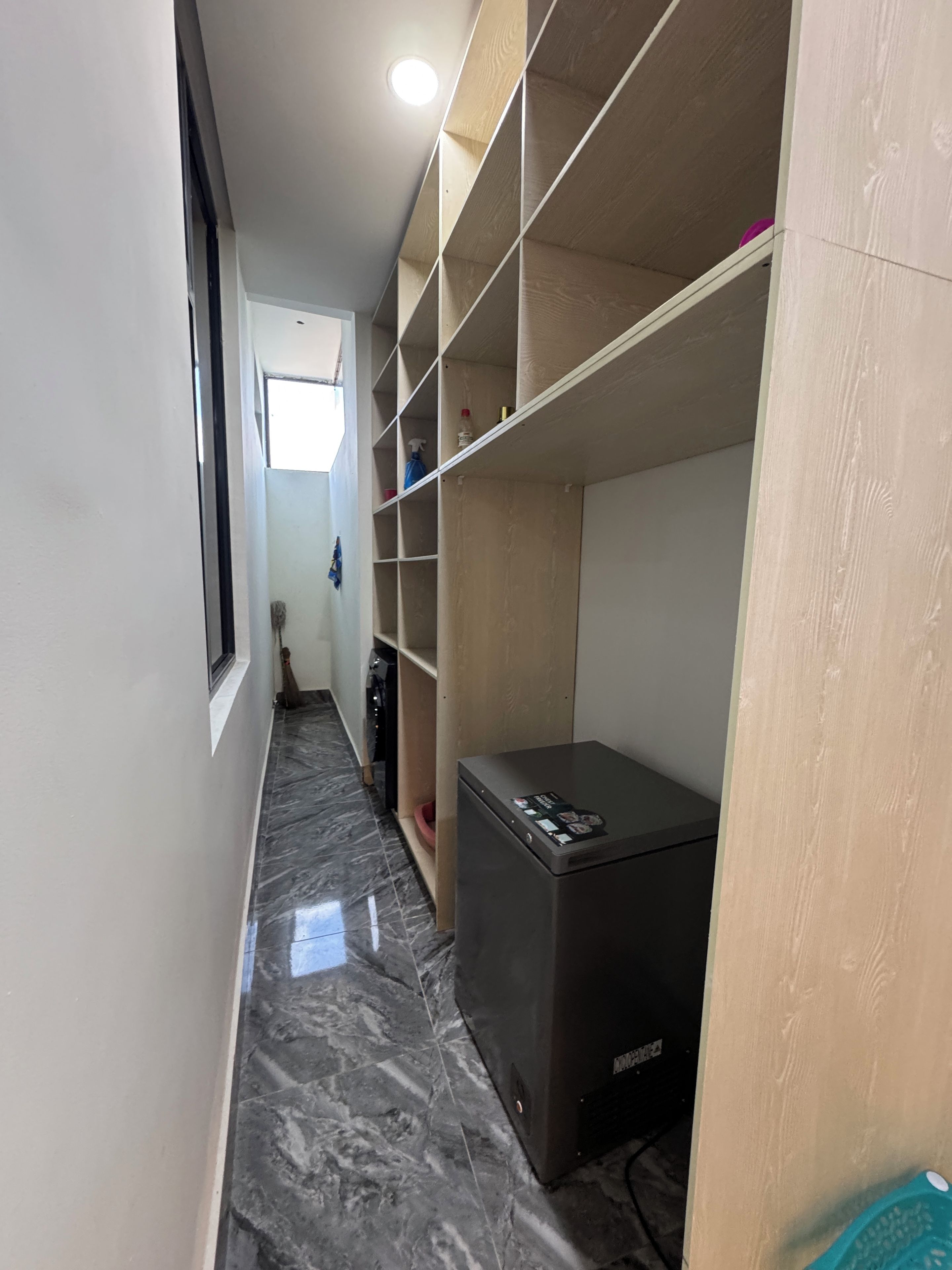 A narrow hallway with a marble floor, a refrigerator, and a wooden shelving unit.
