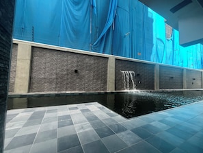 Una piscina con caída de agua, suelo de baldosas y una pared con textura decorativa.