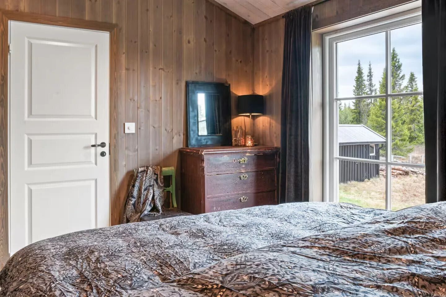 Ein Schlafzimmer mit Holzwand, einem Bett mit gemustertem Überwurf, einem Schminktisch mit Spiegel und einem Fenster mit Blick auf Bäume und ein Gebäude.