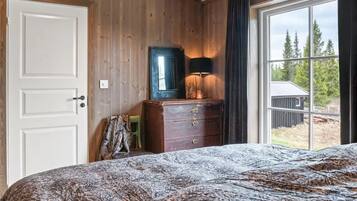 Ein Schlafzimmer mit Holzwand, einem Bett mit gemustertem Überwurf, einem Schminktisch mit Spiegel und einem Fenster mit Blick auf Bäume und ein Gebäude.