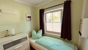 Um quarto pequeno com uma cama de solteiro, cômoda, janela com cortinas e aquecedor na parede.