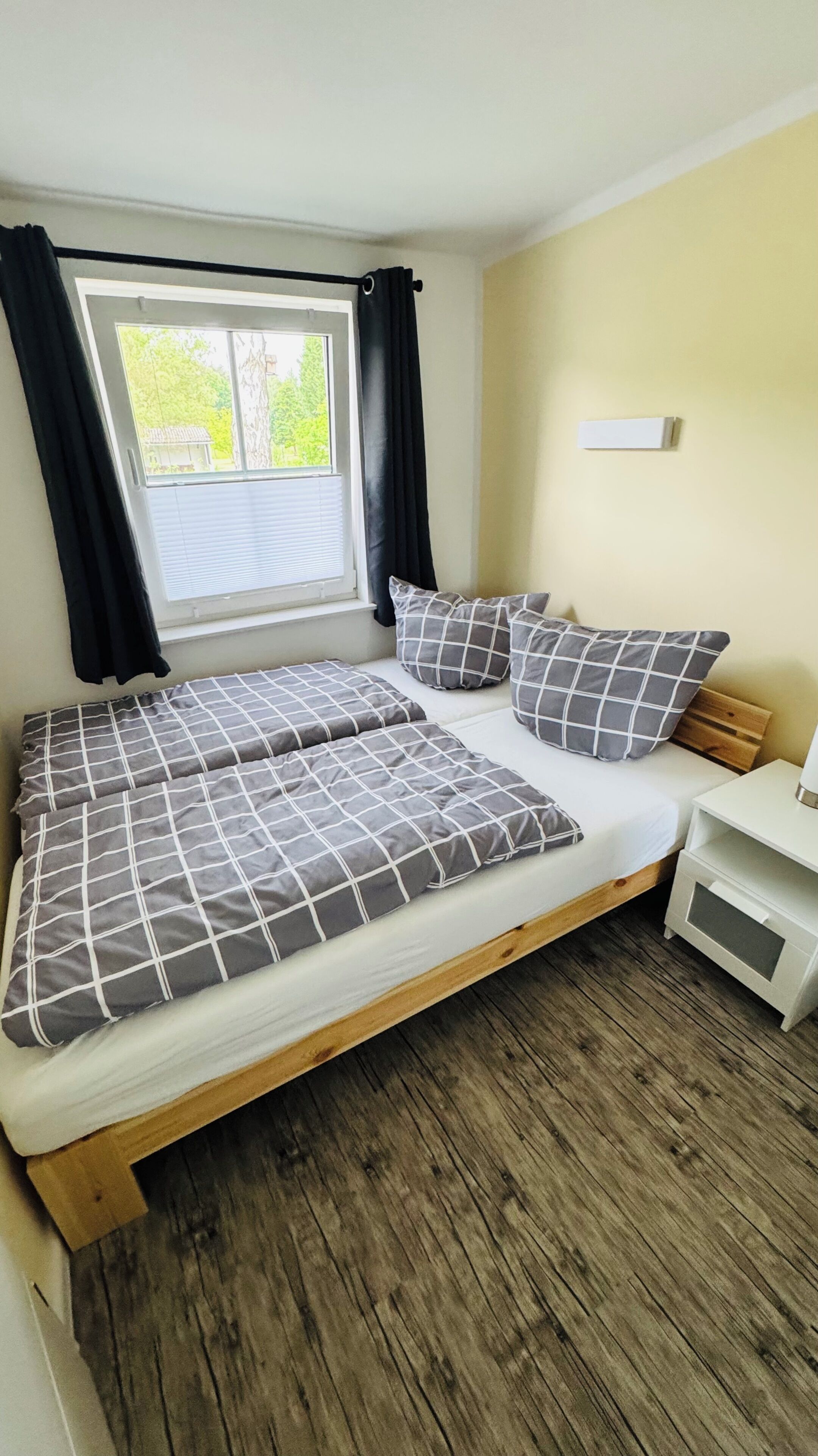 A bed with a wooden frame, a white mattress, and gray checkered bedding. There is a window with black curtains, a bedside table with a lamp, and a small white cabinet.