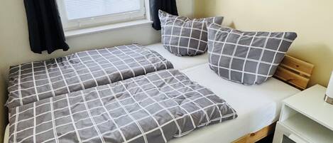 Una cama con estructura de madera, colchón blanco y ropa de cama a cuadros grises. Hay una ventana con cortinas negras, una mesita de noche con lámpara y un mueble blanco pequeño.