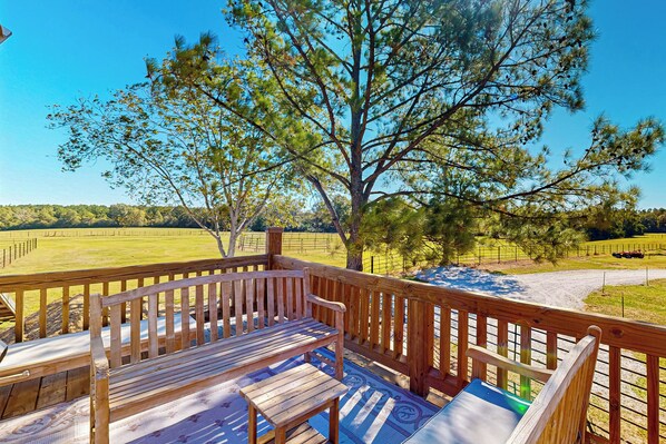 A wooden deck with a bench and a table, overlooking a fenced pasture and a clear blue sky.