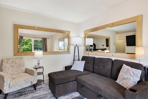 Una habitación con sofá, sillón, dos espejos, una lámpara y vistas a zonas verdes.