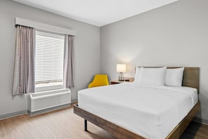 Quarto de hotel com cama, mesa de cabeceira com abajur, uma cadeira e janela com cortinas.