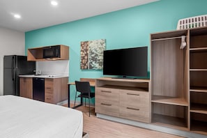 Ein kleines Hotelzimmer mit Bett, Küchenzeile, Flachbildfernseher, Schreibtisch mit Stuhl und Kleiderschrank.