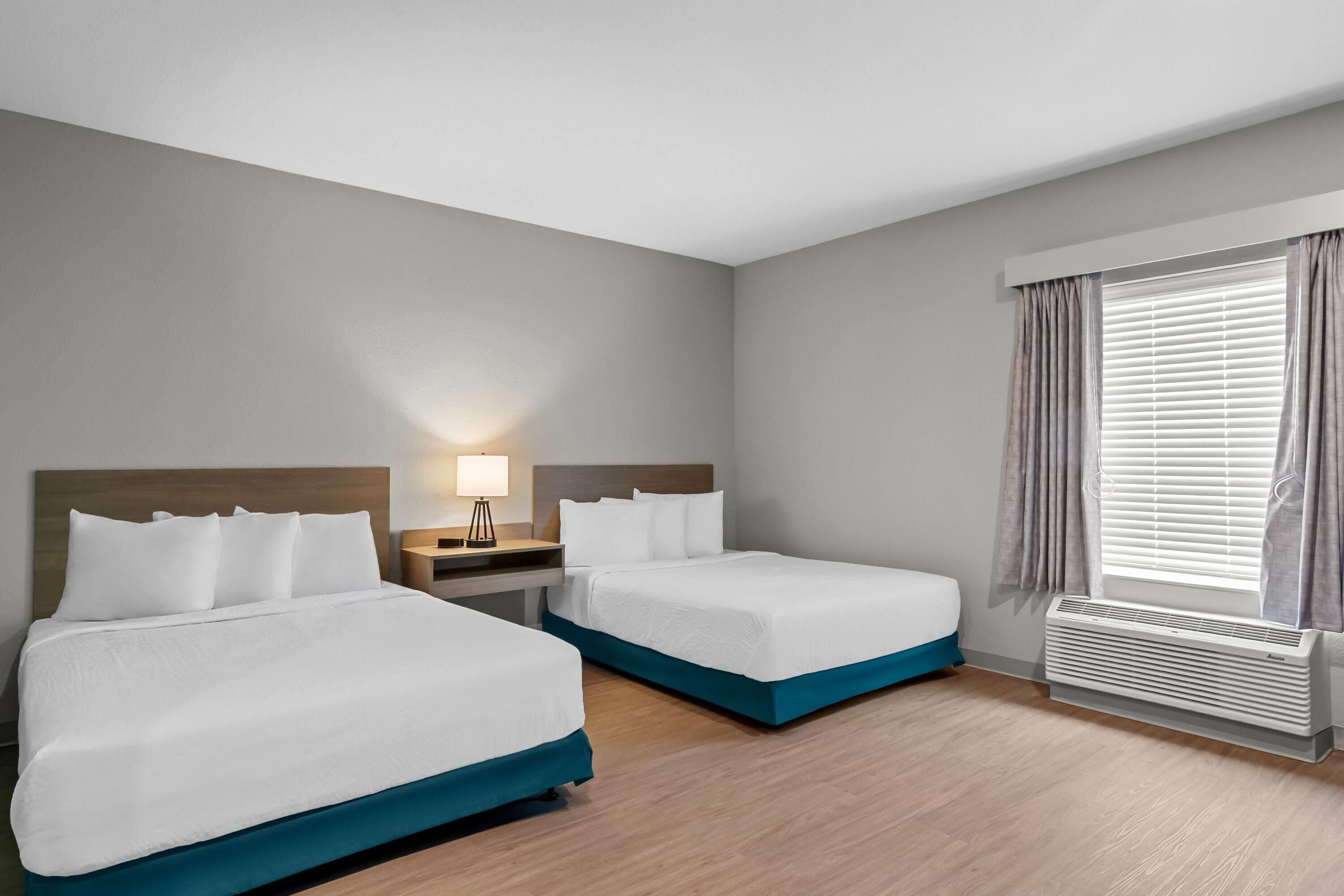 Habitación de hotel con dos camas, una mesita de noche, una lámpara, una ventana con cortinas y un equipo de aire acondicionado.