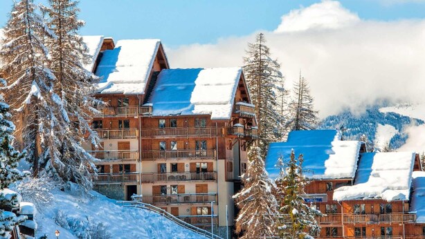 Ein mehrstöckiges Holzchalet mit Balkonen, umgeben von schneebedeckten Bäumen und Bergen.