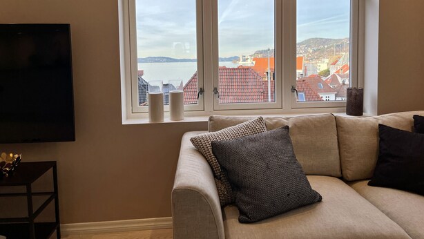 A living room with a beige sofa, a flat-screen TV, and a large window with a view of buildings and water. - Exclusive top apartment, central location in Bergen. (Bergen)
