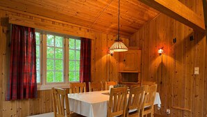 Ein Holzblockhaus-Interieur mit Esstisch, Stühlen, Kamin und einem Fenster mit Vorhängen.