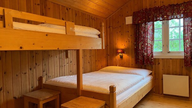 Ein Holzhüttenraum mit Stockbetten, einem Einzelbett, einem kleinen Tisch und einem Fenster mit Vorhängen.