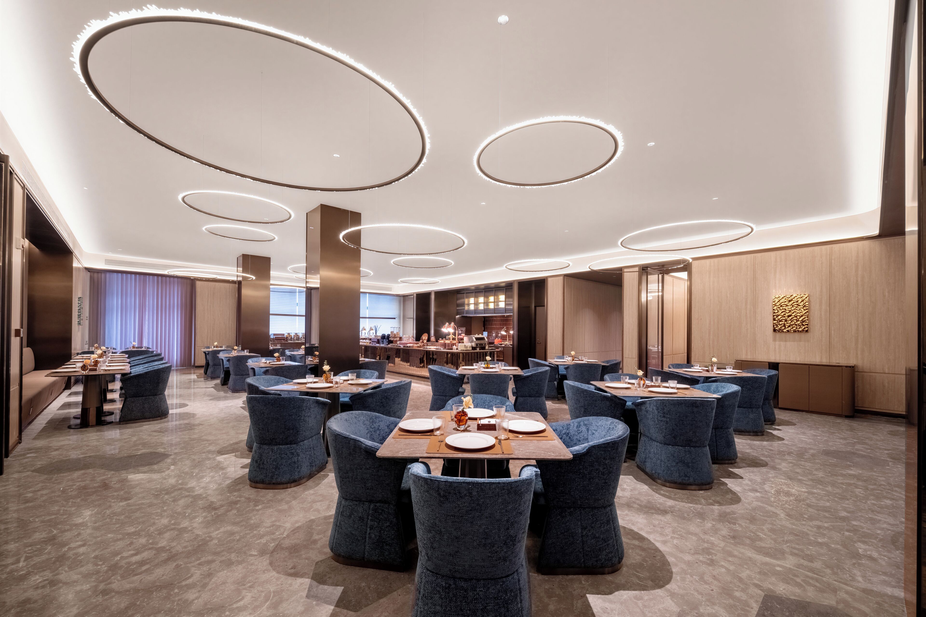 A modern hotel dining area with round tables, blue upholstered chairs, and circular ceiling lights.