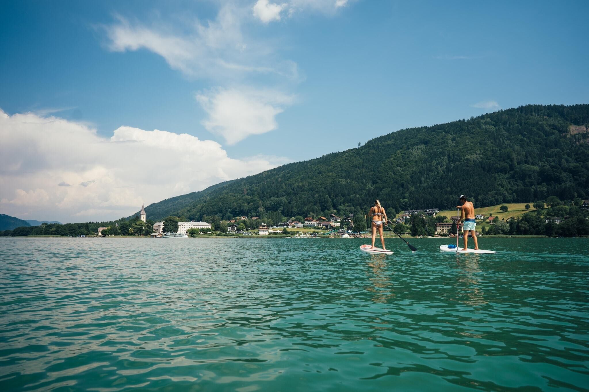 A lake with two paddleboarders, a church, and a hill in the background.