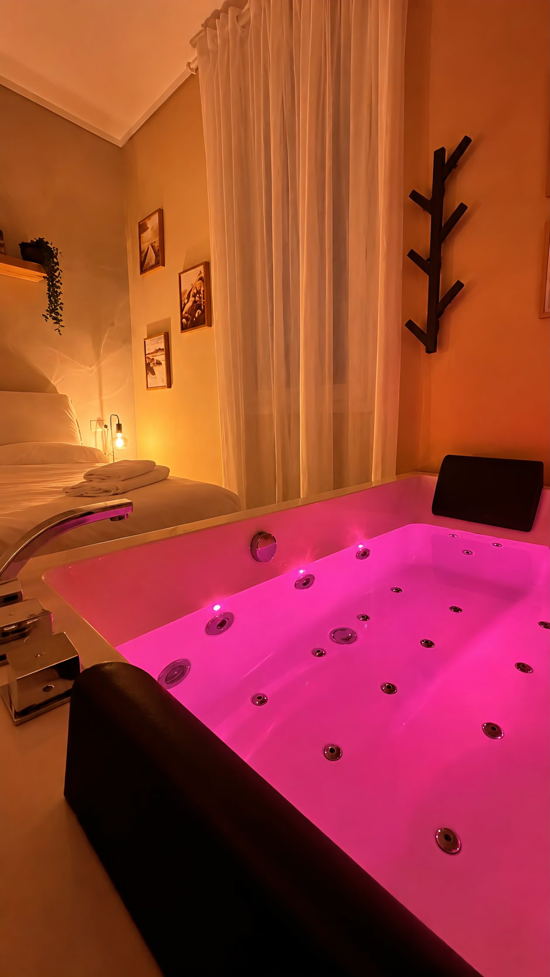A room with a large pink hot tub, a bed with white linens, a wall-mounted coat rack, and framed pictures.