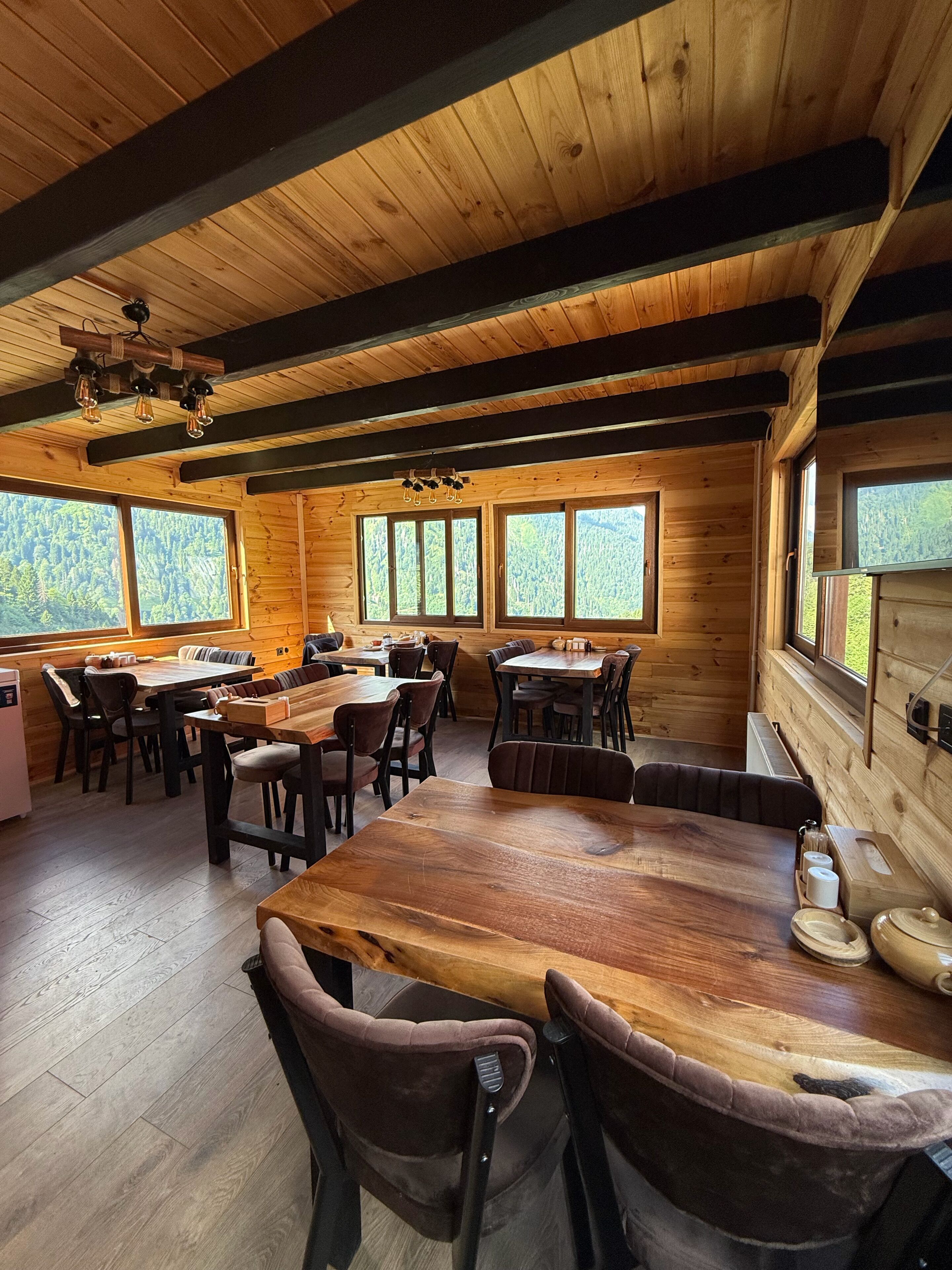 A wooden cabin interior with a large dining table, multiple chairs, and large windows offering a view of the forest.