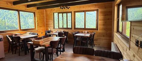 A wooden cabin interior with a large dining table, multiple chairs, and large windows offering a view of the forest.