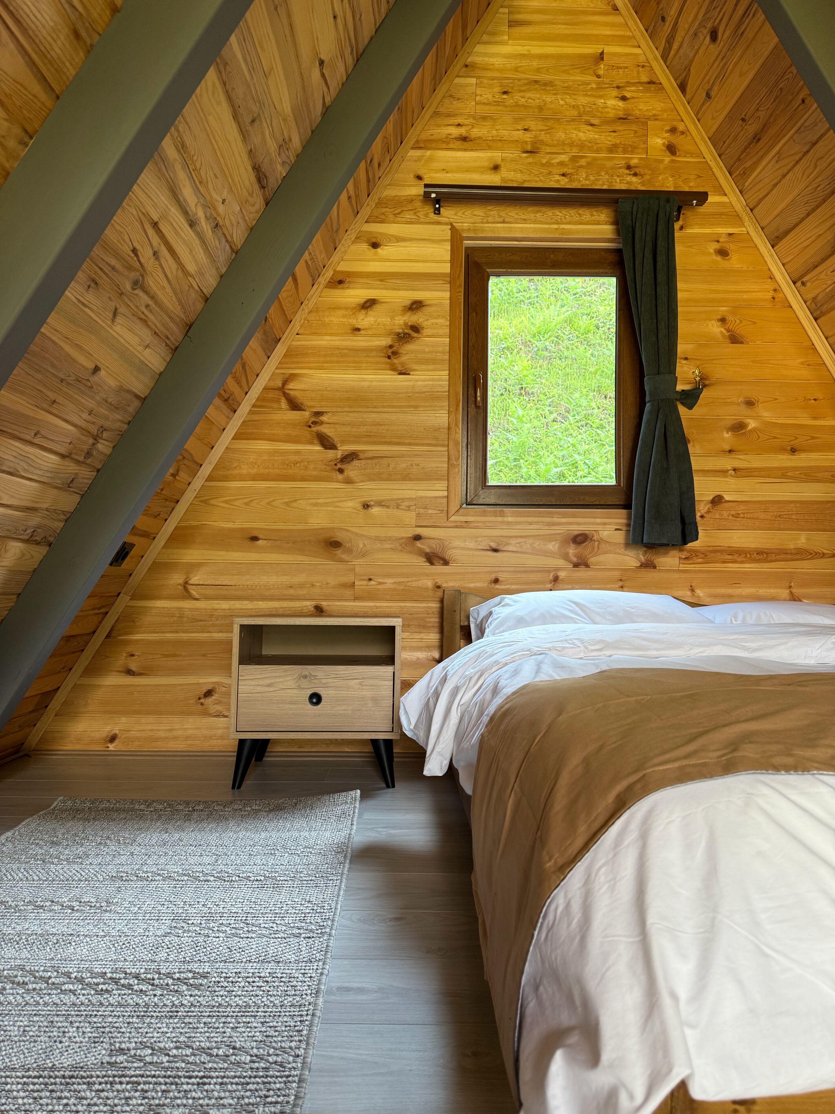 A bedroom with a sloped ceiling, a window with curtains, a bed, a nightstand, and a rug.