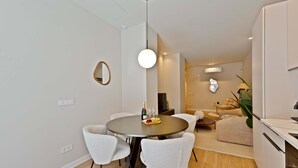 Een moderne eetruimte met een ronde tafel, witte stoelen, een hanglamp en uitzicht op een woonruimte met een bank en een plant.