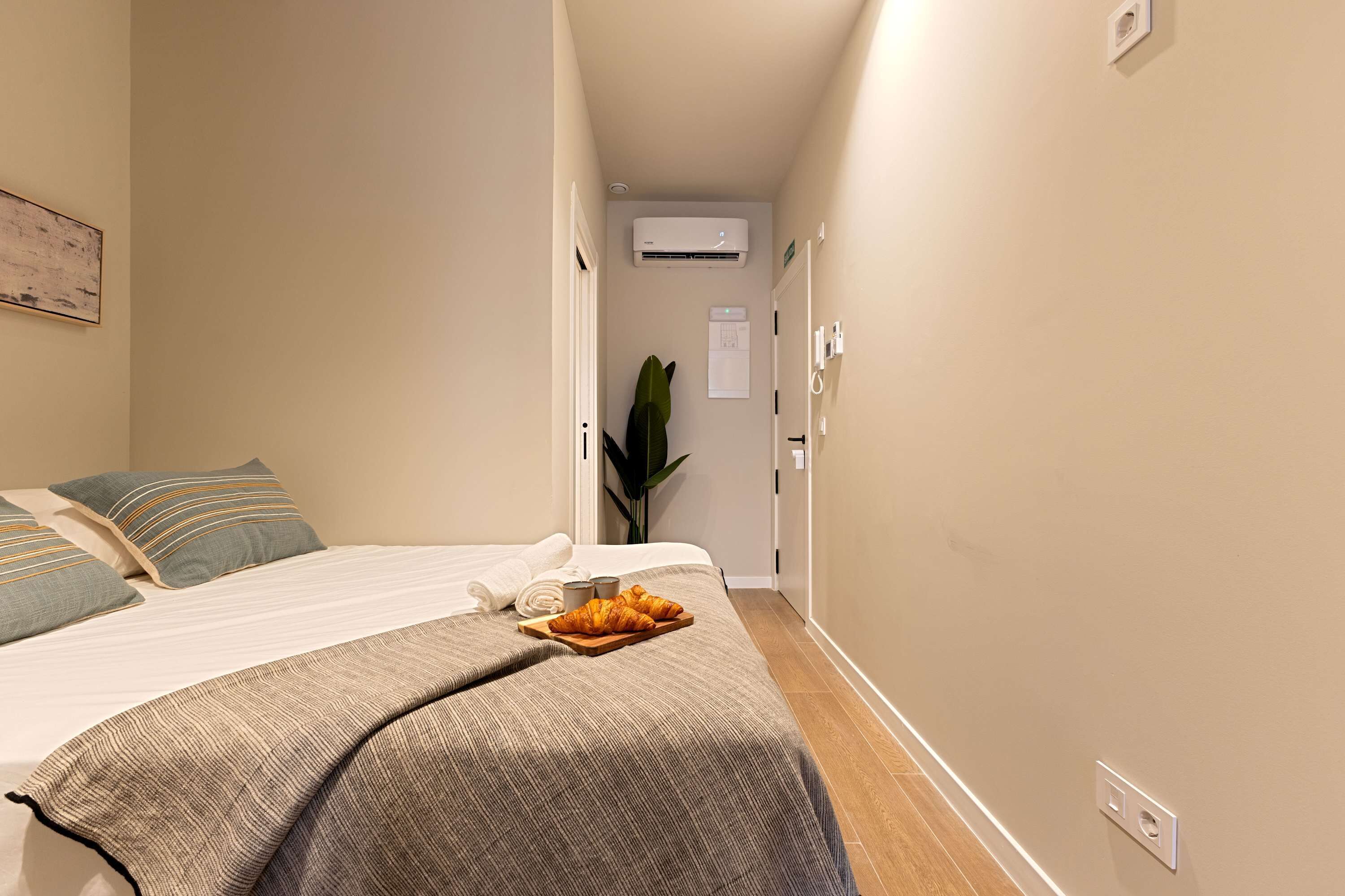 A hotel room with a bed, a bedside table with a tray of croissants, a plant, and a wall-mounted air conditioner.