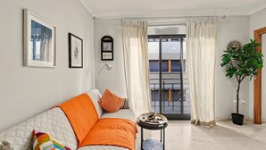 Een woonkamer met een bank, een klein bijzettafeltje, een potplant, een raam met gordijnen en een deur.