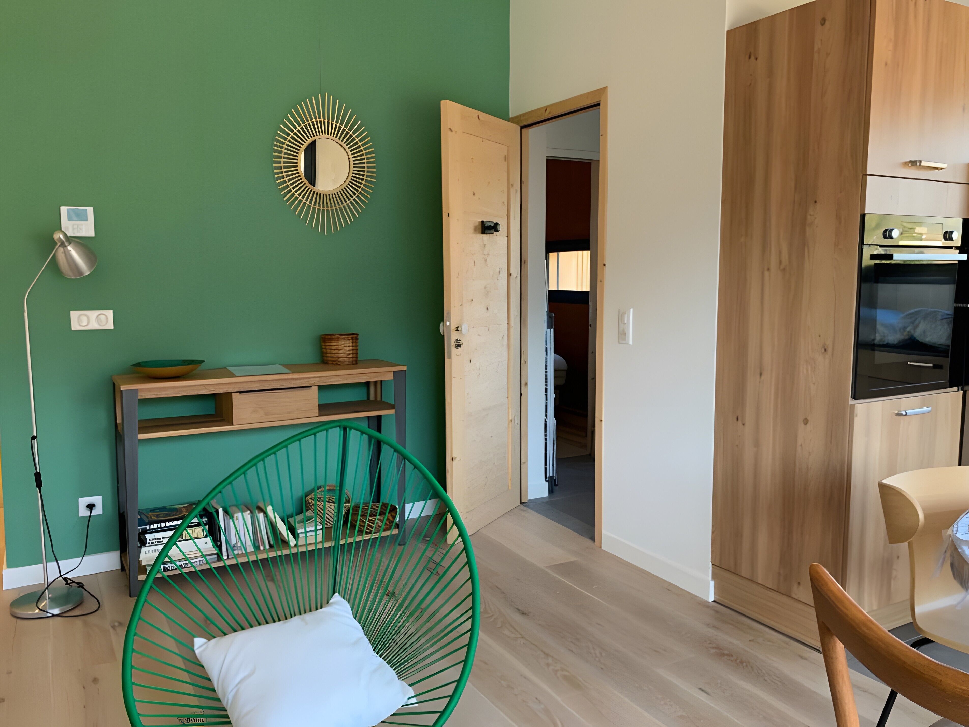 Una habitación con una pared verde, un armario de madera, una silla verde, suelo de madera y un espejo.