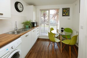 A compact kitchen with a washing machine, a sink, a clock, a window with blinds, and a dining area with chairs.
