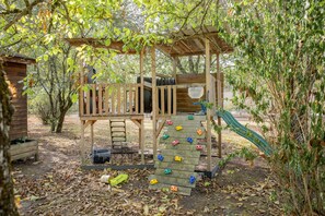 A wooden play structure with a slide and climbing wall, surrounded by trees and a shed.