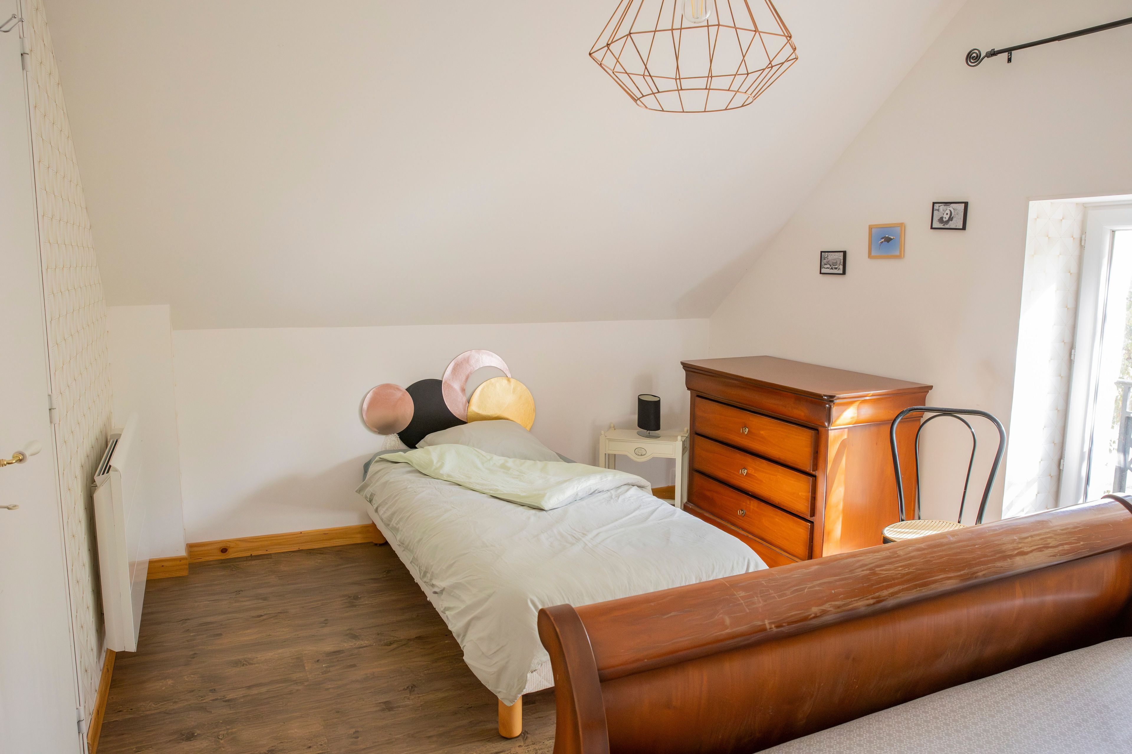 A bedroom with a bed, a wooden dresser, a bedside table with a lamp, and a hanging light fixture.