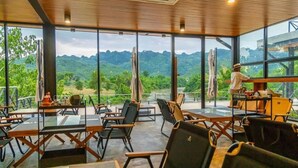 A restaurant with large windows offering a view of green hills. Tables and chairs are arranged for dining.