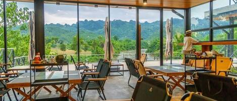 A restaurant with large windows offering a view of green hills. Tables and chairs are arranged for dining.