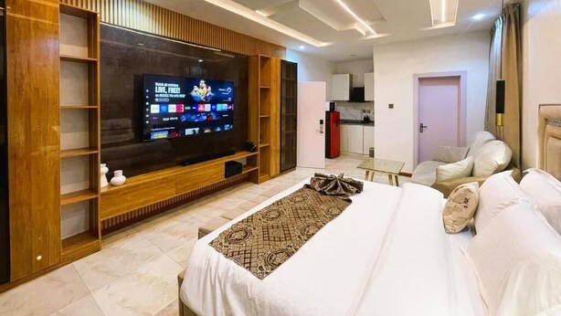 A modern hotel room with a large flat-screen TV, a bed with white linens, a wooden entertainment unit, and a kitchenette in the background. - Waveline Apartment (Abuja)