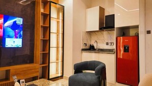A modern living room with a flat-screen TV, a wooden shelving unit, a grey armchair, a glass coffee table, and a red refrigerator. - Waveline Apartment (Abuja)