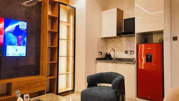 Een moderne woonkamer met een flatscreen televisie, een houten wandplank, een grijze fauteuil, een glazen salontafel en een rode koelkast.