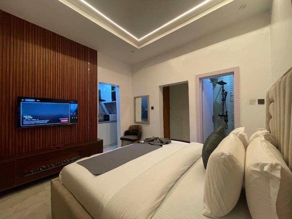 Une chambre à coucher moderne, avec un lit, un téléviseur fixé sur un panneau en bois, et une salle de bain visible à travers une porte ouverte.