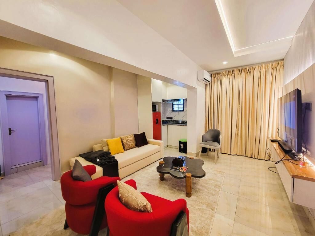 A modern living room with a sofa, red chairs, a coffee table, a TV, and a kitchen area visible through an open door.