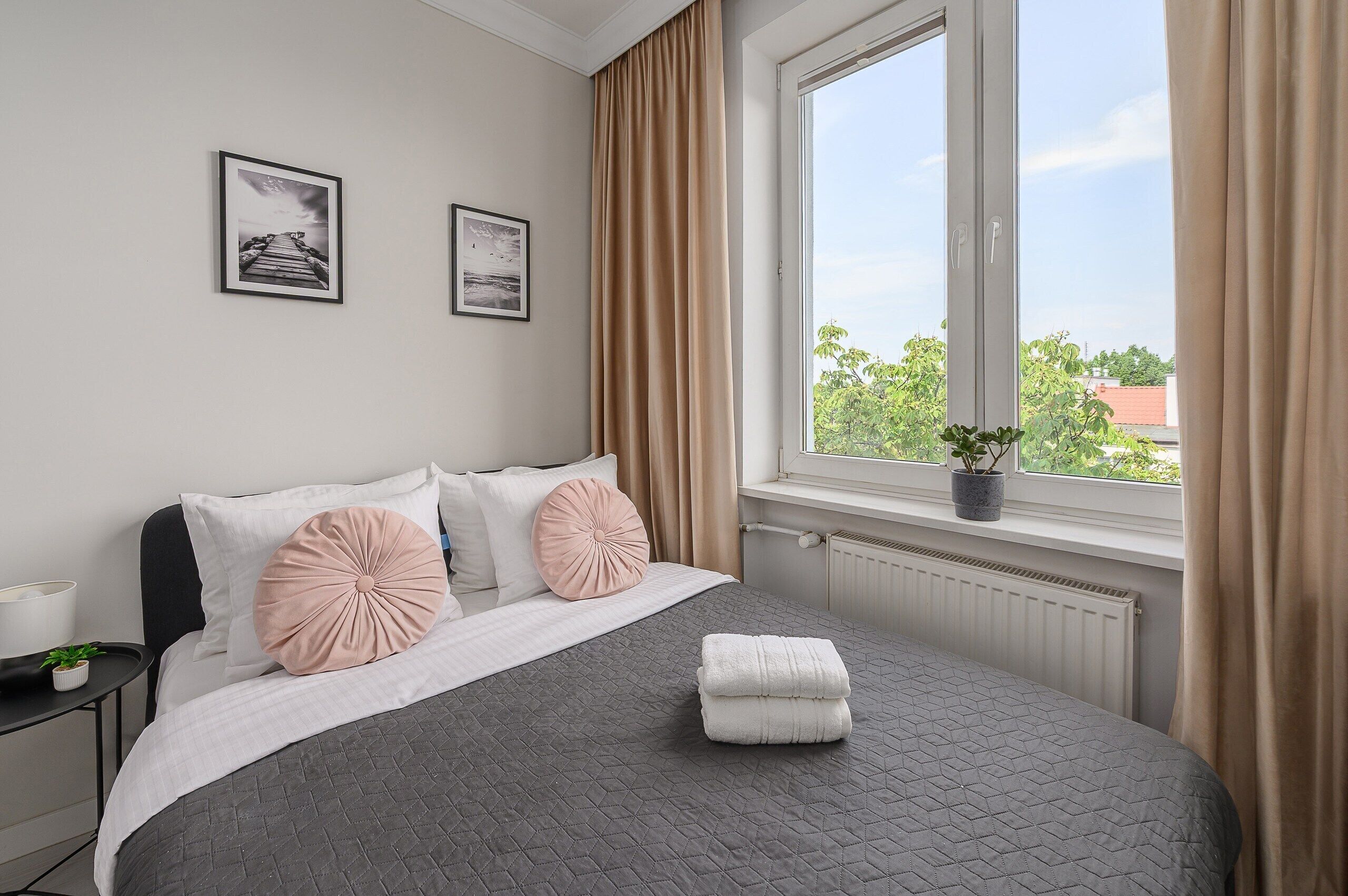 Ein ordentlich eingerichtetes Schlafzimmer mit einem Bett, Kissen, einem Nachttisch mit Lampe, einem Fenster mit Vorhängen und Blick ins Grüne.