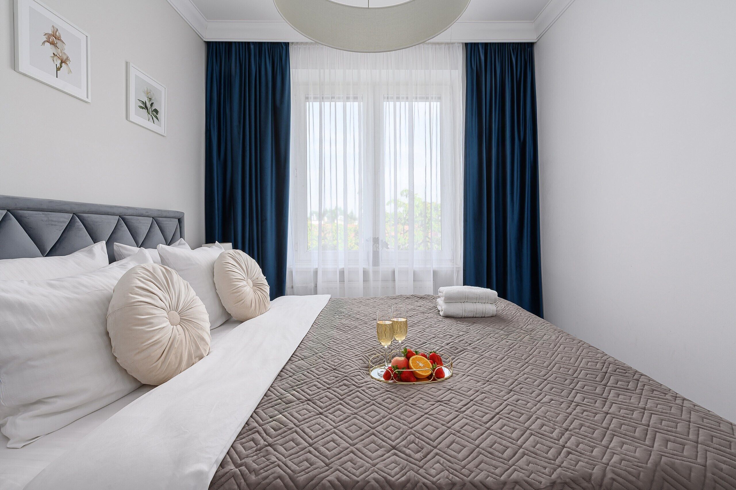 Ein Hotelzimmer mit einem Bett, einer Ablage mit Früchten und zwei Sektgläsern, sowie Blick durch durchscheinende Vorhänge.