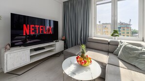 Un salon moderne doté d’un téléviseur à écran plat, d’un canapé, d’une table basse avec des fruits, et offrant une vue sur les immeubles à travers de grandes fenêtres.