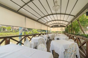 An outdoor covered dining area with tables set for guests, a wooden deck railing, and a view of greenery and buildings in the background. - Room 'Villa Goetzen - Camera Singola' with Wi-Fi and Air Conditioning (Dolo)