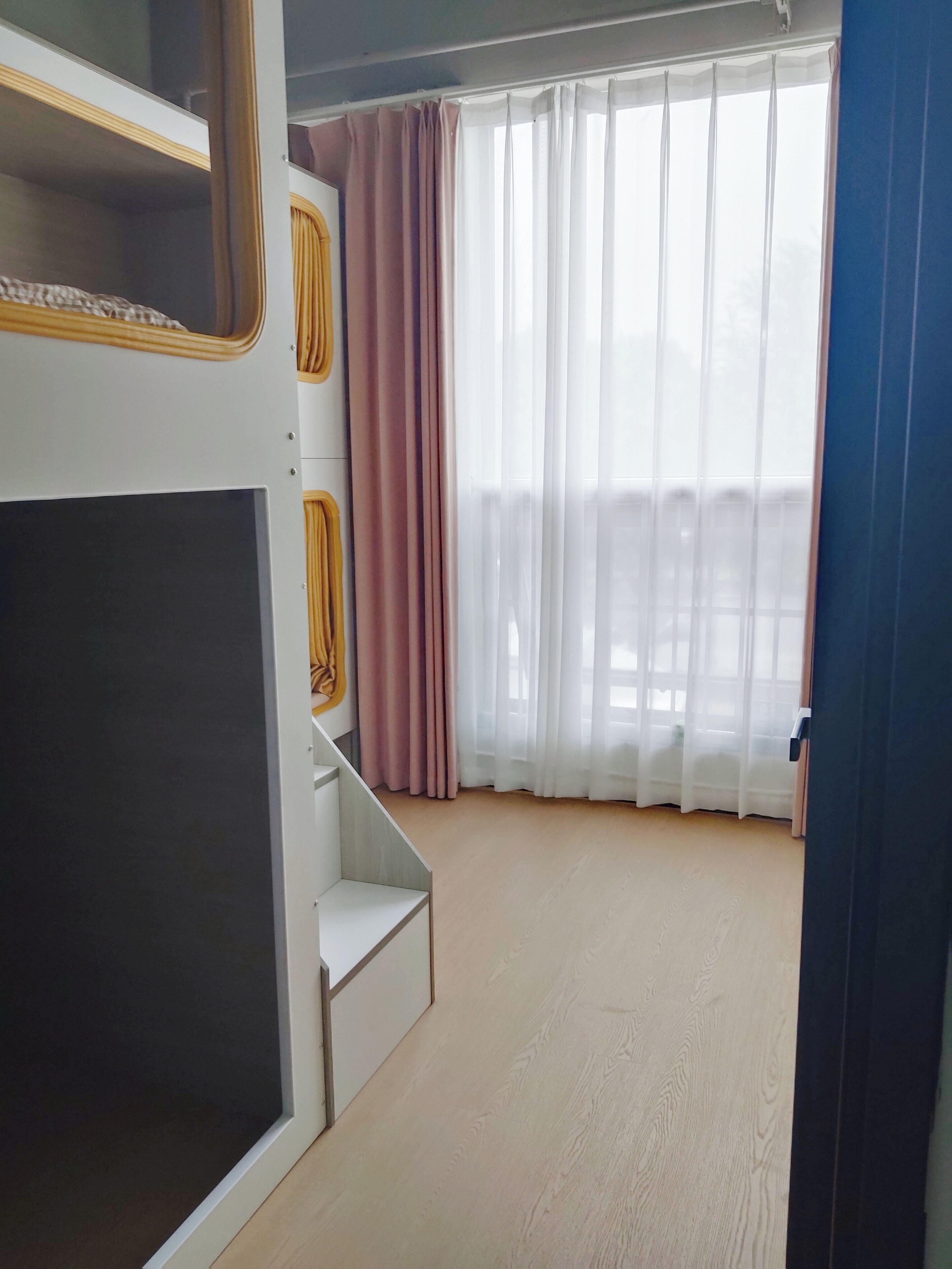 A room with a bunk bed, a small staircase, a large window with sheer curtains, and a view of the outside.