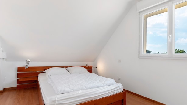 A bedroom with a wooden bed, white bedding, a bedside table, and a window with a view of the sky.