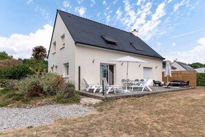 A modern two-story house with a sloped roof, a patio with outdoor furniture, and a garden area.
