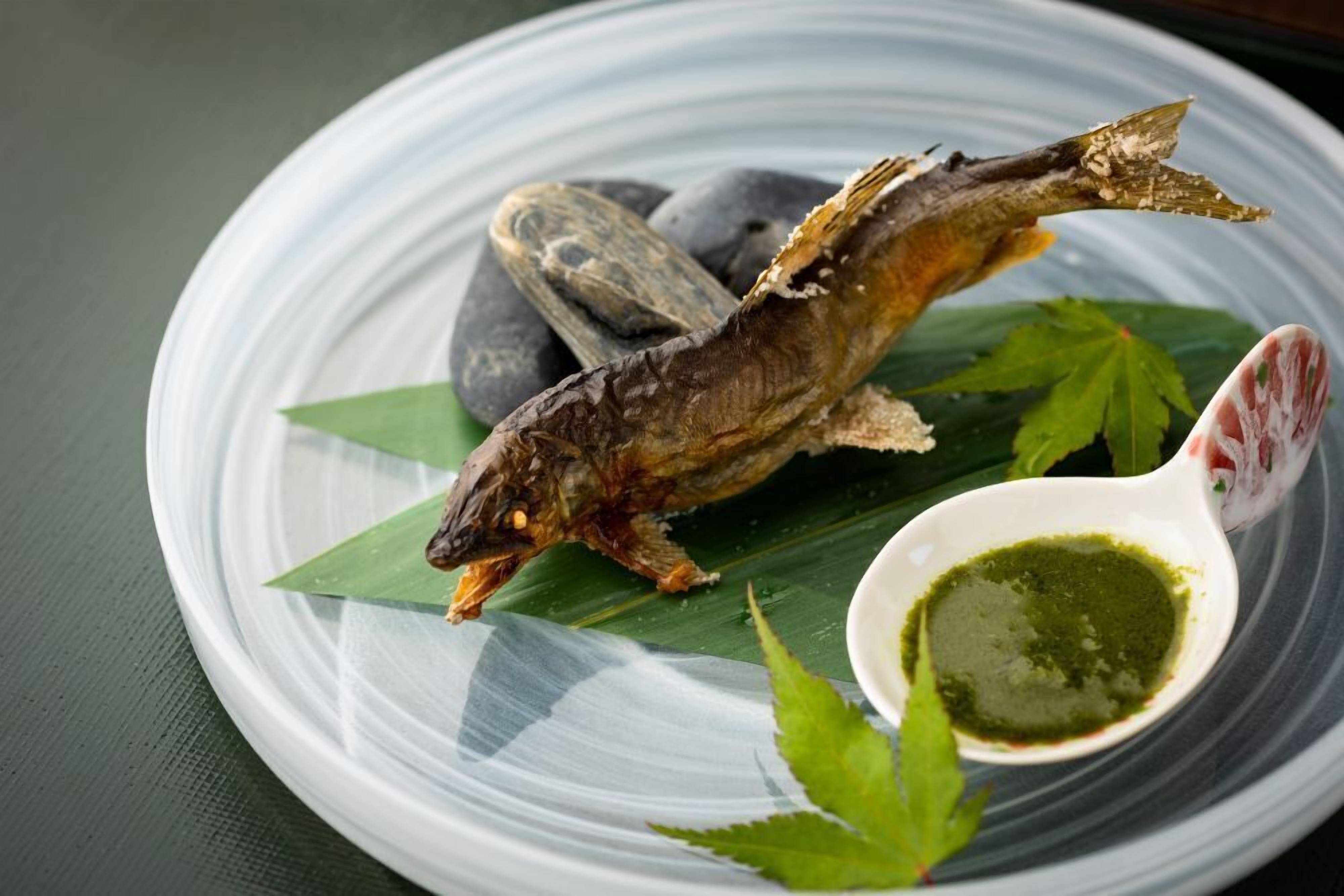 Un plato con un pescado entero, algunas hojas verdes y un recipiente pequeño con salsa verde.