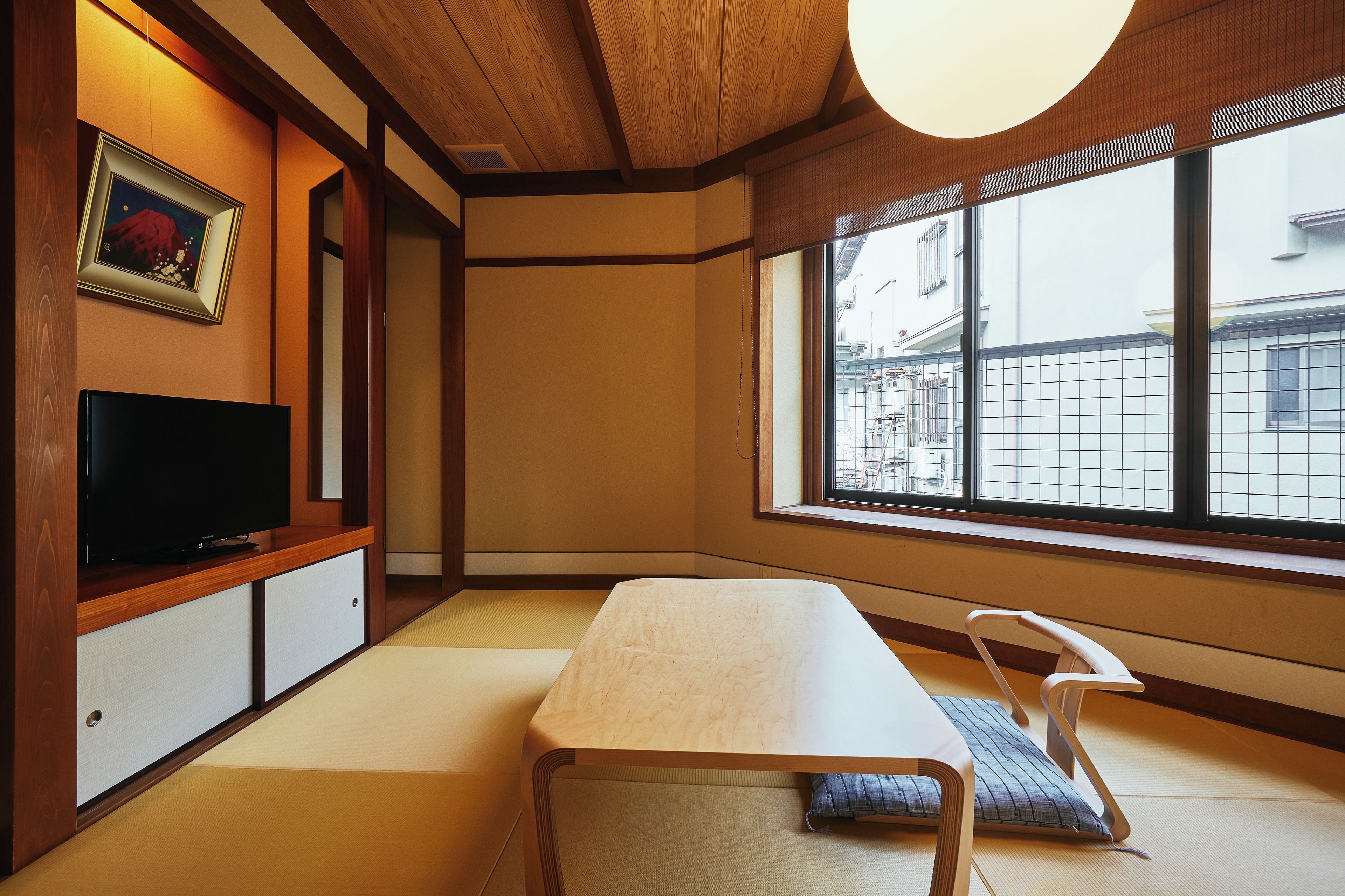 Una habitación japonesa tradicional con suelo de tatami, una mesa baja, un televisor y una ventana con vistas a edificios.
