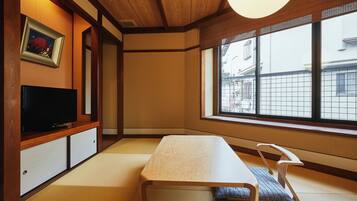 Una habitación japonesa tradicional con suelo de tatami, una mesa baja, un televisor y una ventana con vistas a edificios.