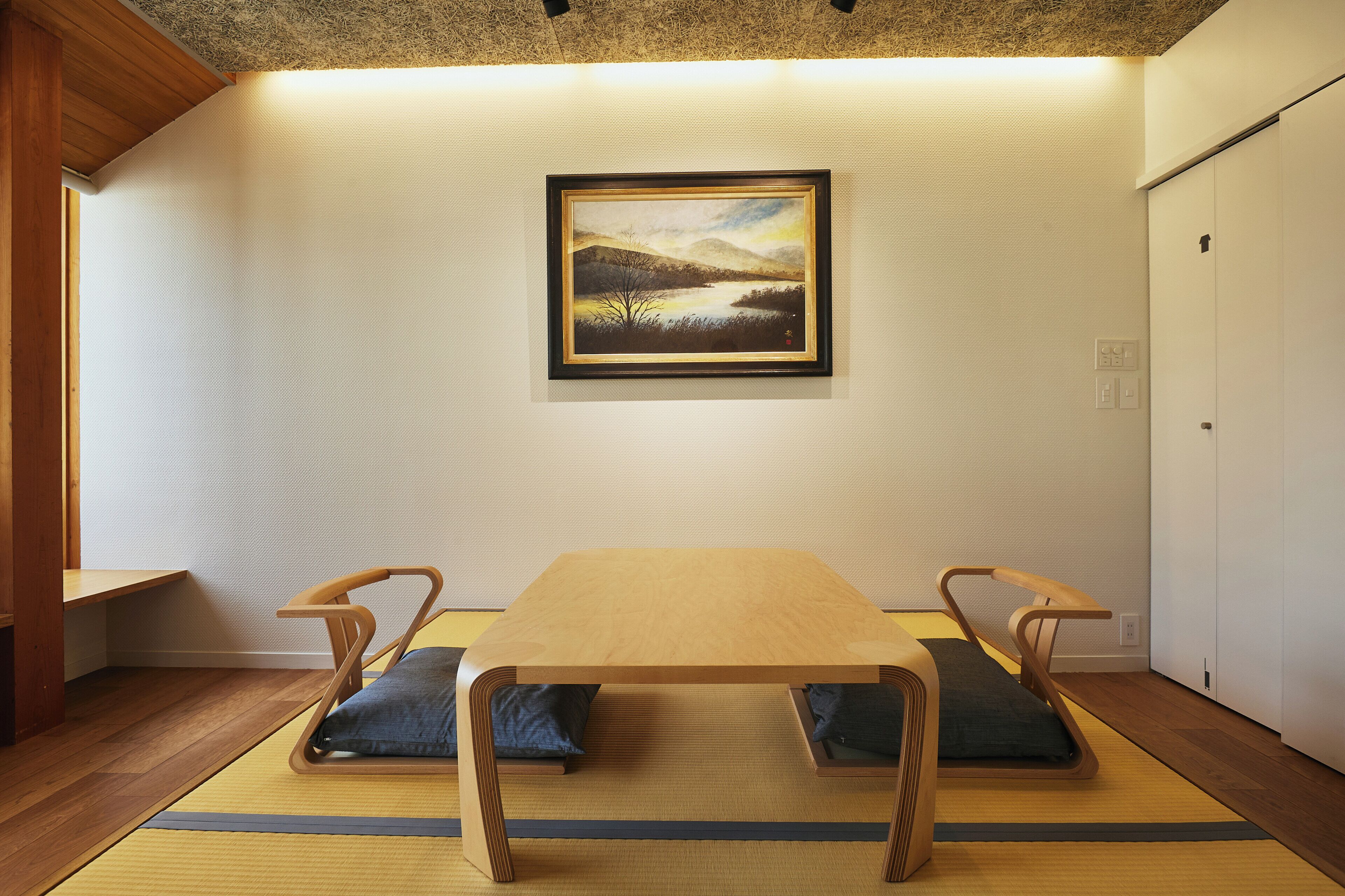 Quarto com mesa de madeira, duas cadeiras, esteiras de tatame, um quadro emoldurado e uma porta branca.