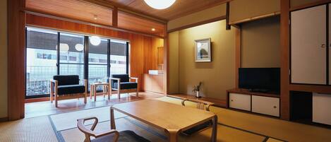 A traditional Japanese-style room with tatami flooring, a low table, chairs, a television, and a window with blinds.