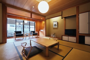A traditional Japanese-style room with tatami flooring, a low table, chairs, a television, and a window with blinds. - Kinosaki Onsen TSUCHIYA  (Toyooka)